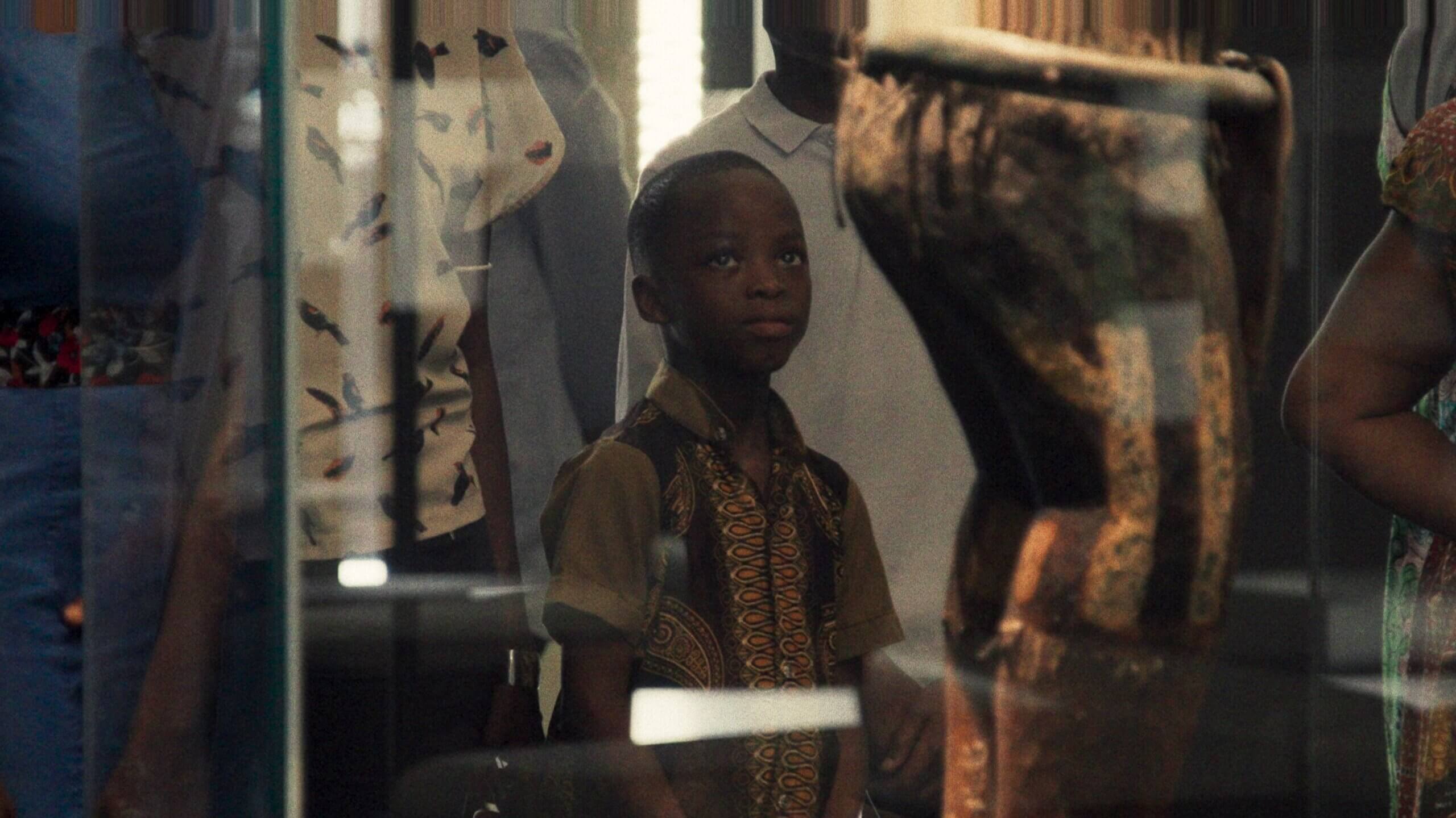 Un jeune béninois regarde un trésor royal de Dahomey derrière une vitrine dans une scène du film Dahomey.