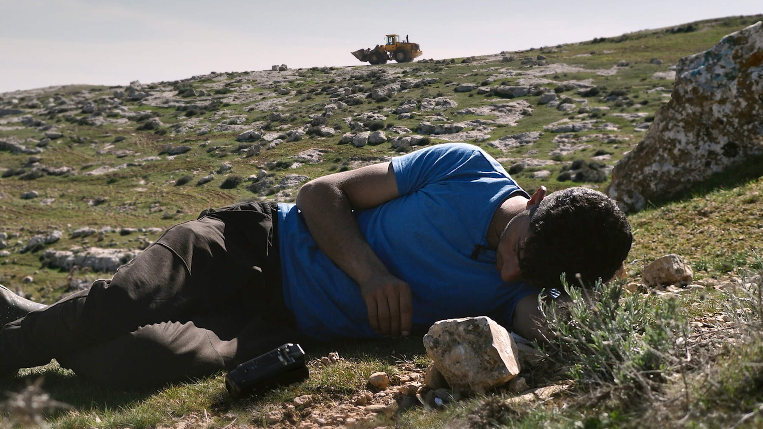 Un jeune homme couché dans l'herbe. Derrière lui, au loin, un bulldozer. Image tirée du film No Other Land.