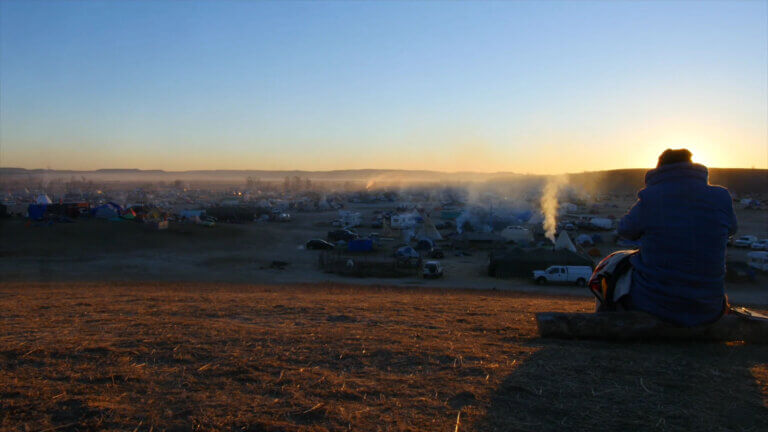 Un homme assis, vu de dos, regarde le camp Standing Rock dans le film Dislocation Blues de Sky Hopinka.