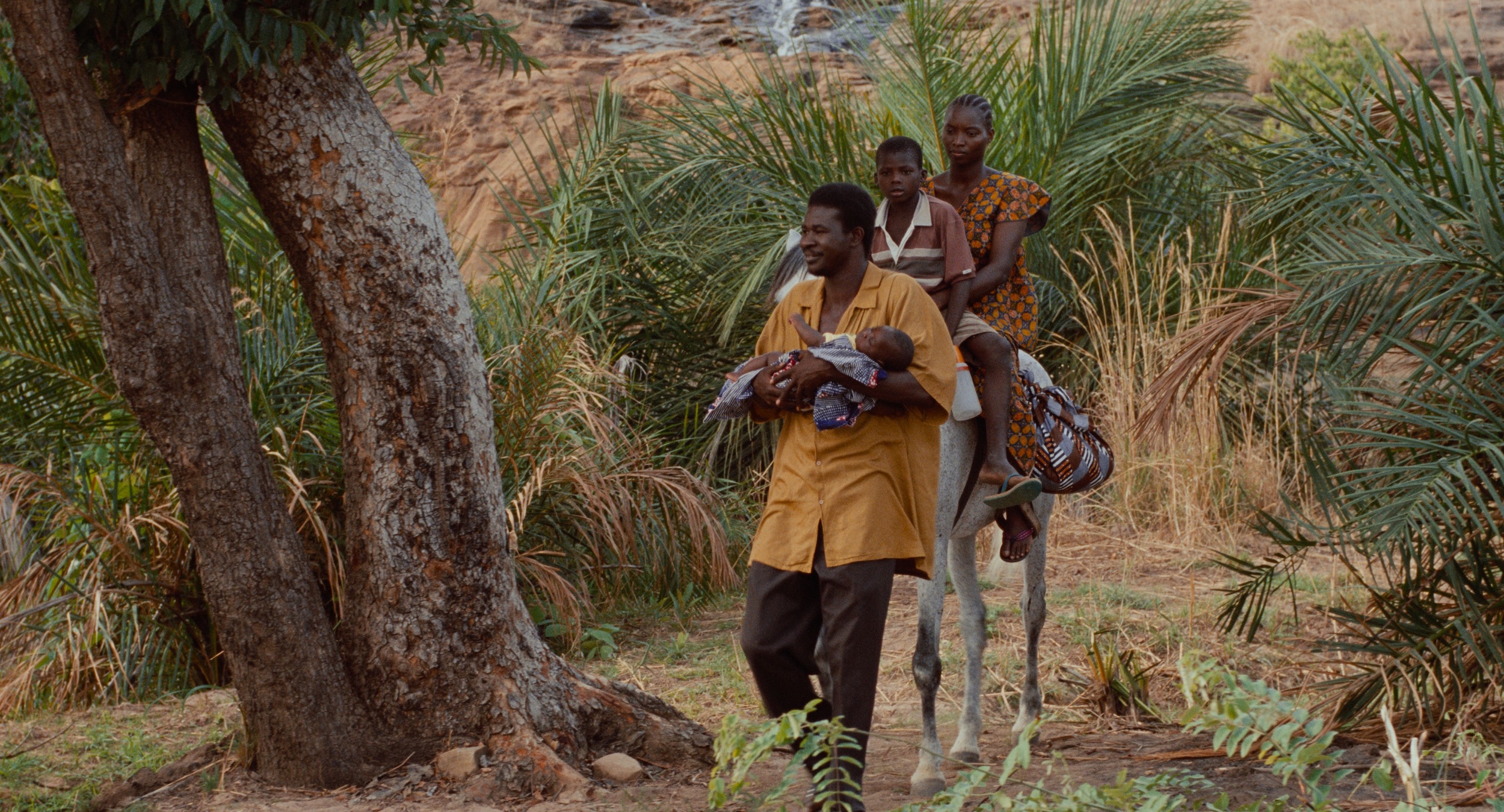 Parmi les palmiers au Burkina Faso, un homme tient un bébé dans ses bras, suivi d'une femme et d'un enfant à cheval.